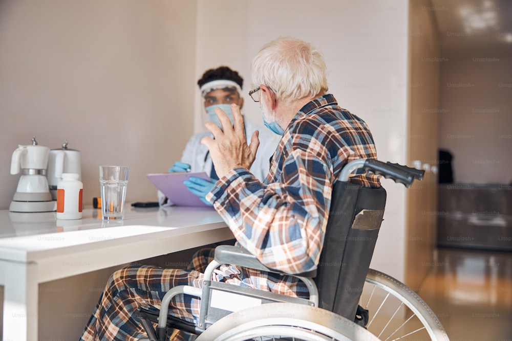 Uomo caucasico anziano su una sedia a rotelle che parla con un giovane medico in una maschera facciale