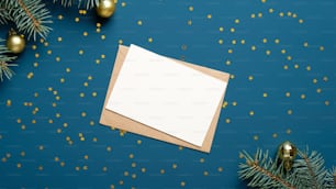 Blank card and craft paper envelope on blue background decorated with confetti and fir branches. Holiday invitation mockup.
