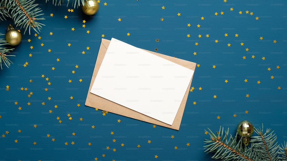 紙吹雪とモミの枝で飾られた青い背景に白紙のカードとクラフト紙の封筒。休日の招待状のモックアップ。