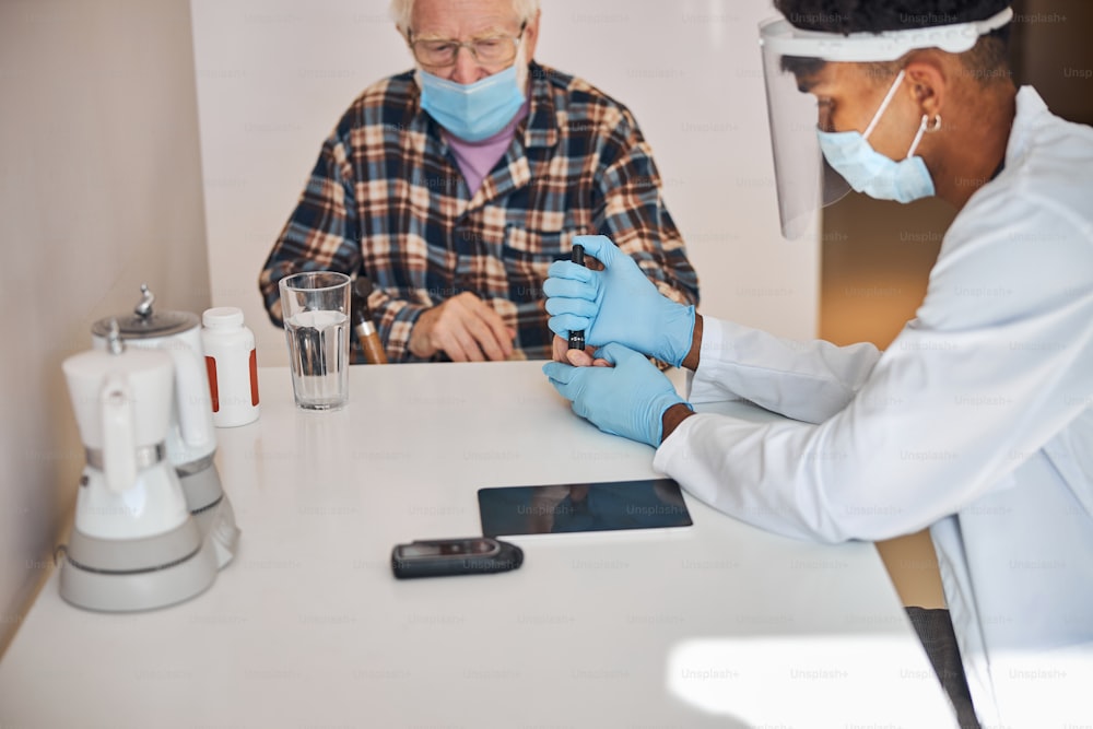 Focused young doctor in latex gloves pricking the senior man fingertip with a lancing device