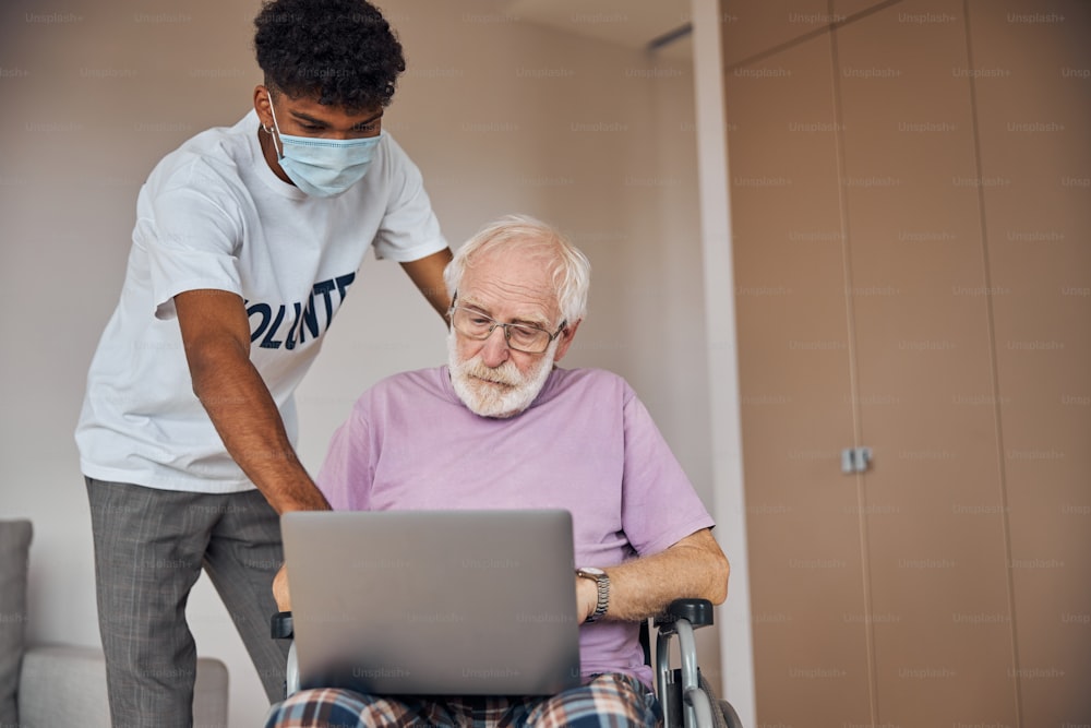 Jovem em uma máscara médica de proteção instruindo o homem com deficiência a usar seu laptop