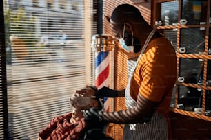 의료 마스크를 쓴 아프리카계 미국인 남성 미용사는 고객 얼굴에서 원치 않는 머리카락을 제거하면서 직선 면도기를 사용합니다.