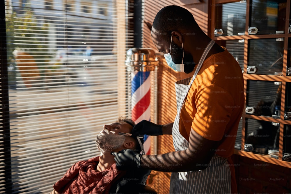 Afro American male hairdresser in medical mask using straight razor while removing unwanted hair from client face