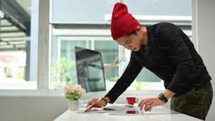 집중된 젊은 남성 디자이너는 컴퓨터와 함께 테이블에 서서 홈 오피스에서 프로젝트를 진행하는 동안 컬러 샘플이 있는 종이를 검사합니다.
