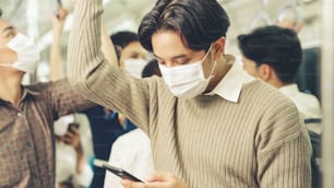 Traveler wearing face mask while using mobile phone on public train . Coronavirus disease or COVID 19 pandemic outbreak and urban city lifestyle problem in rush hour commuting concept .