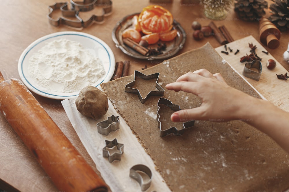 Hände schneiden Lebkuchenteig mit festlichen Stern- und Baummetallschneidern auf rustikalem Tisch mit Gewürzen, festlicher Dekoration, Lichtern. Person, die Weihnachtslebkuchenplätzchen herstellt, Feiertagsadvent