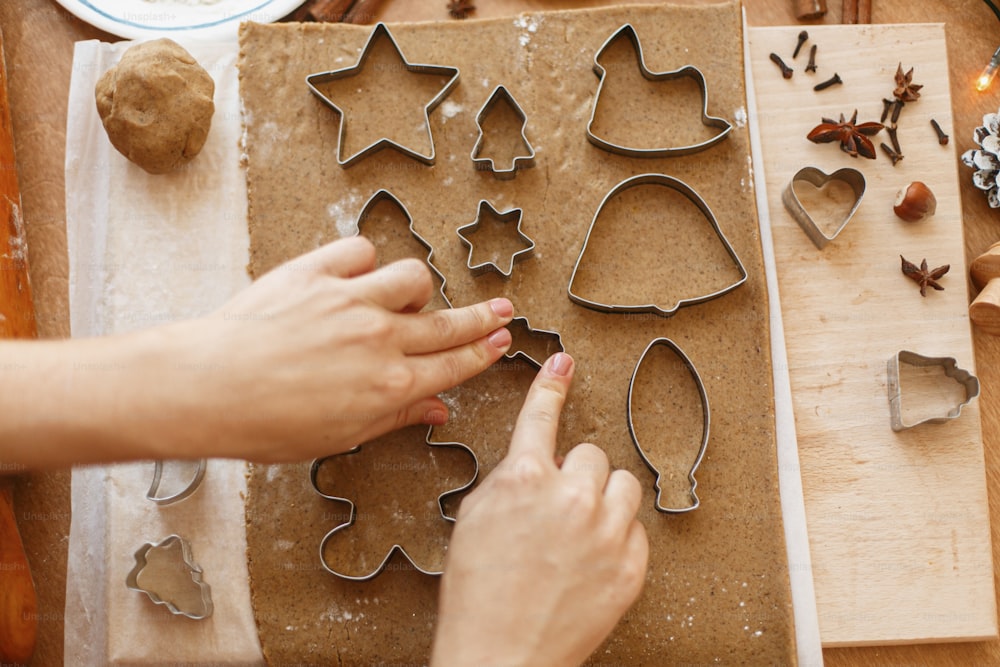Person making Christmas gingerbread cookies, holiday advent. Hands cutting gingerbread dough with festive metal cutters on rustic table with spices, oranges, festive decorations, rolling pin