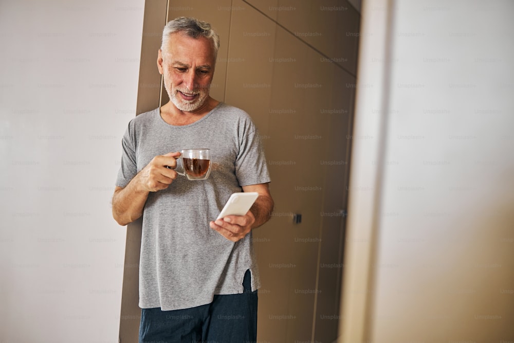 Anciano relajado con ropa cómoda sonriendo mientras sostiene una taza de té y su teléfono inteligente