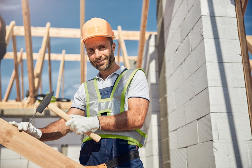 Gentil constructeur barbu gardant le sourire sur son visage tout en travaillant sur un grand chantier de construction