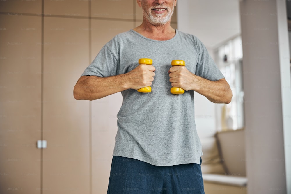 Cropped photo of a grey-haired man holding a pair of dumb-bells close to his chest while working out
