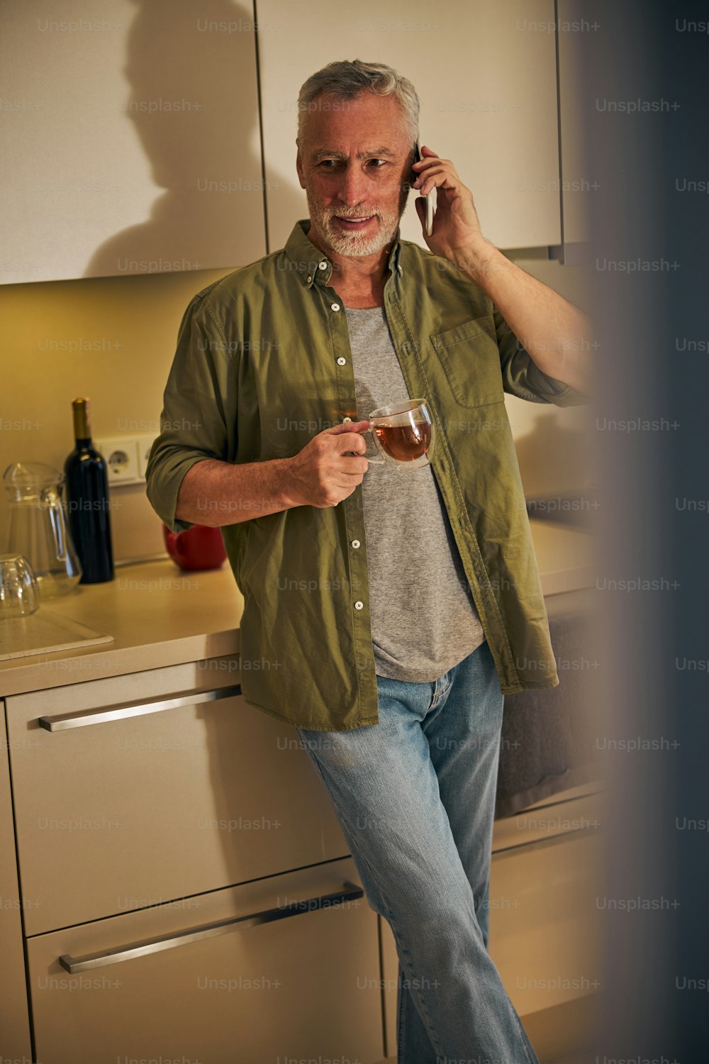Uomo anziano che parla al telefono mentre tiene in mano una tazza di tè e si trova nella sua cucina