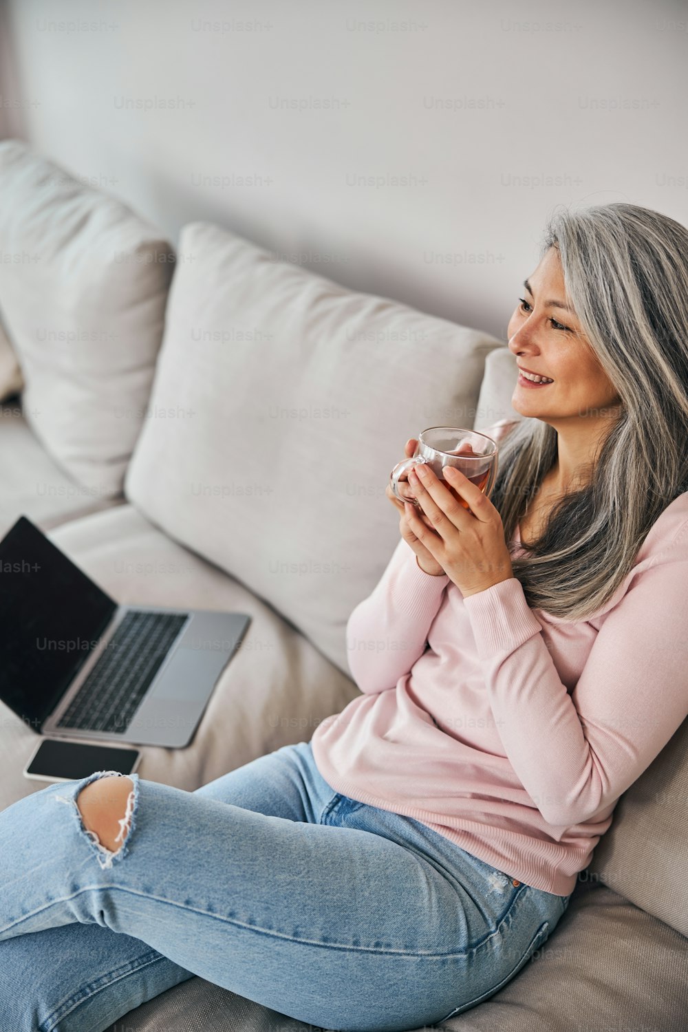 Beautiful lady holding cup of hot drink and smiling while resting on comfortable couch with laptop