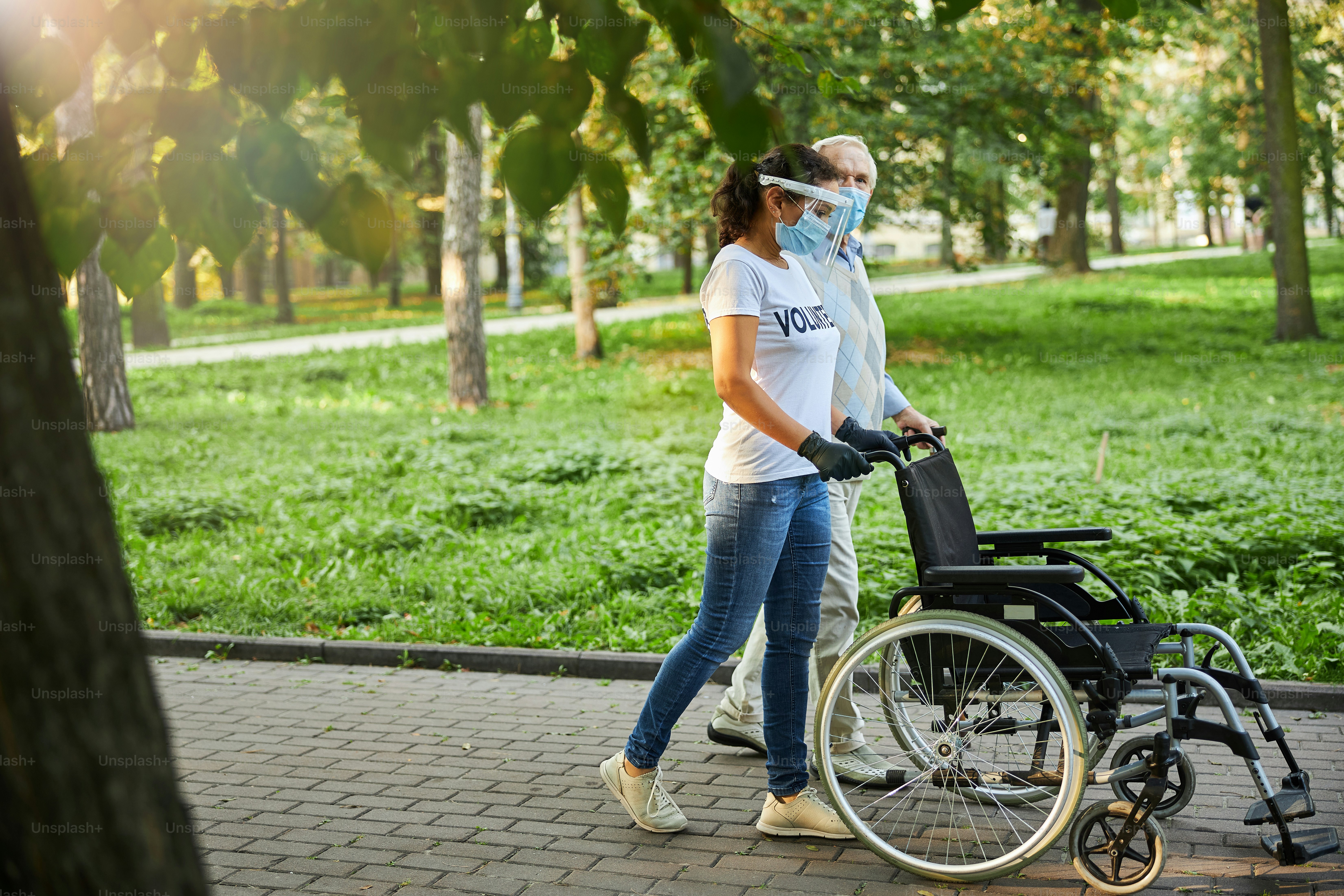 Side view portrait of social worker and older man walking in the city park or wood with wheelchair