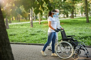 Side view portrait of social worker and older man walking in the city park or wood with wheelchair