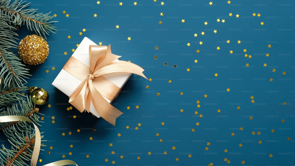 Christmas gift box decorated ribbon bow, fir tree branches, golden confetti blue background. Christmas and New Year banner. Flat lay, top view, copy space.