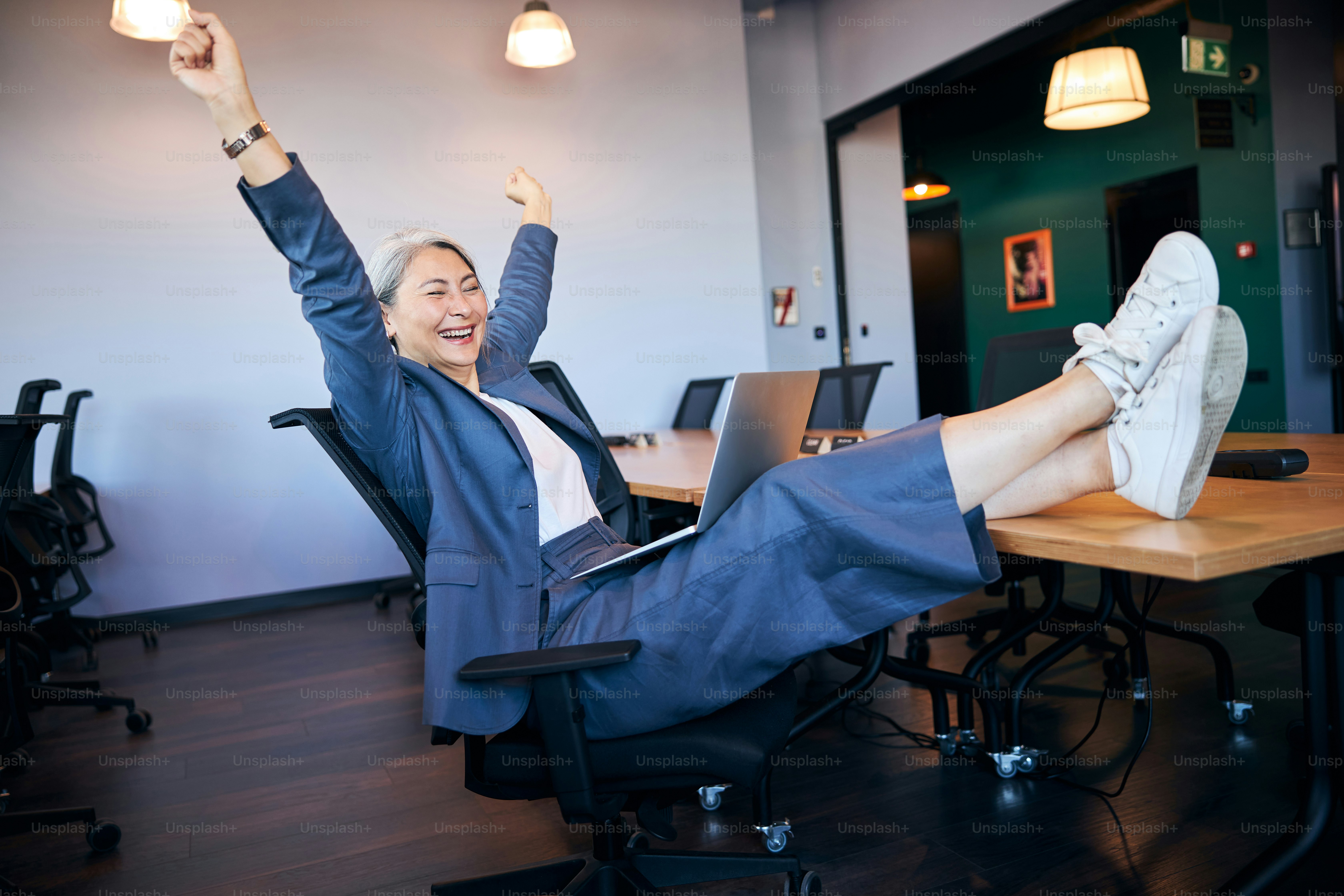Joyful businesswoman with notebook on her laps raising hands and smiling while sitting in chair and placing her feet on desk