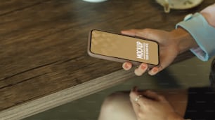 Colpo ritagliato di mano femminile che tiene lo smartphone sul tavolo di legno, include il percorso di ritaglio