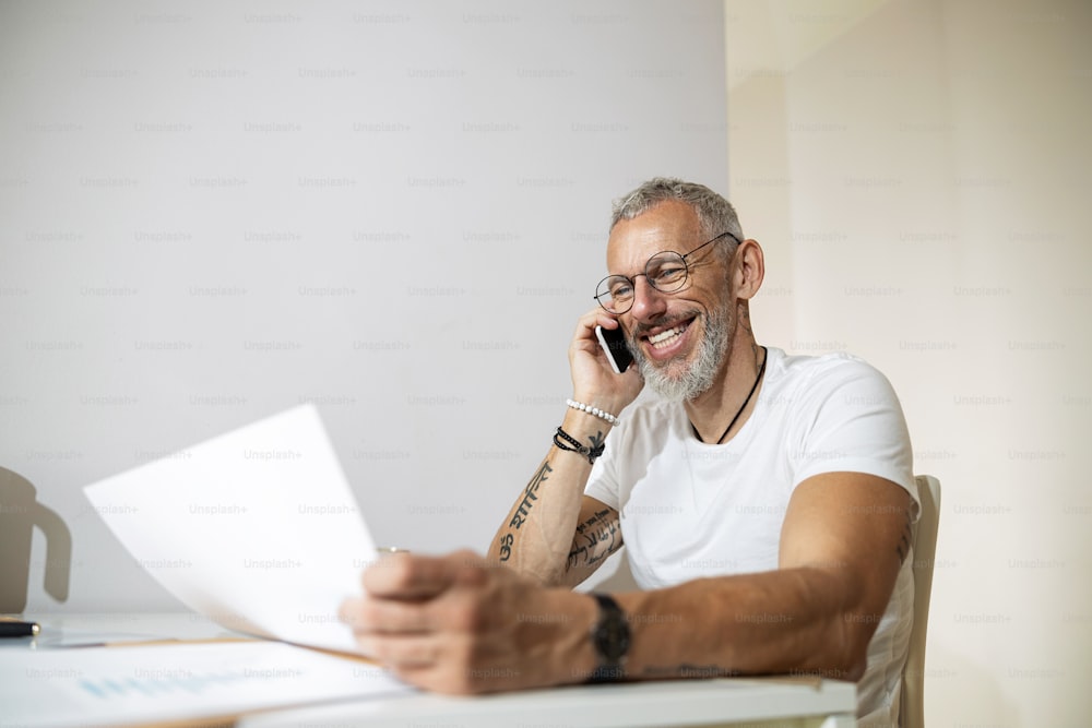 Homme d’affaires heureux à lunettes, pressant un smartphone à son oreille tout en discutant et en vérifiant une feuille de papier