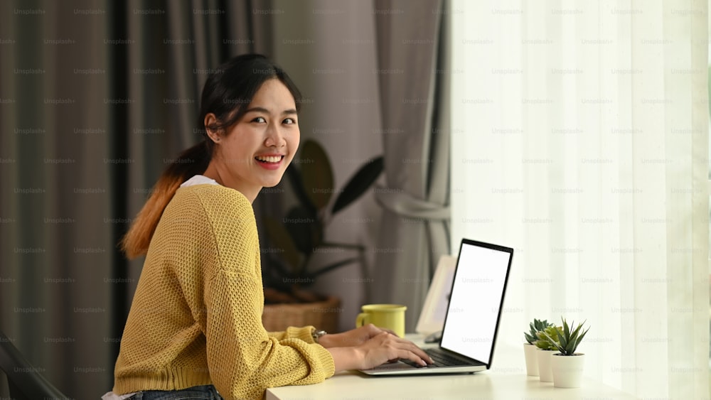 A young female freelancer working on laptop while sitting at her workspace and smiling to camera.