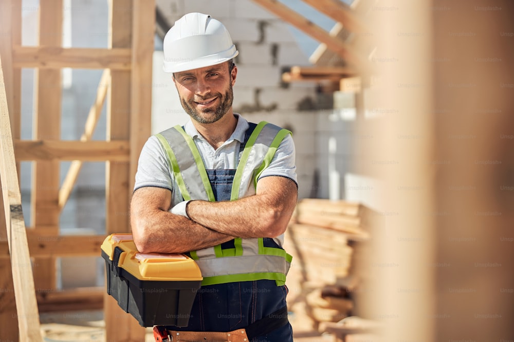 Joven constructor alegre que usa casco mientras trabaja en el sitio de construcción, sosteniendo el caso con el instrumento