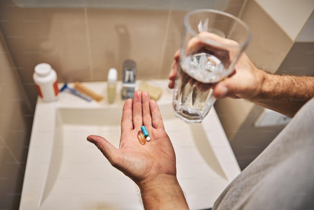 Abgeschnittenes Foto von Händen, die ein Glas Wasser und einen Haufen Pillen mit einem Waschbecken im Hintergrund halten