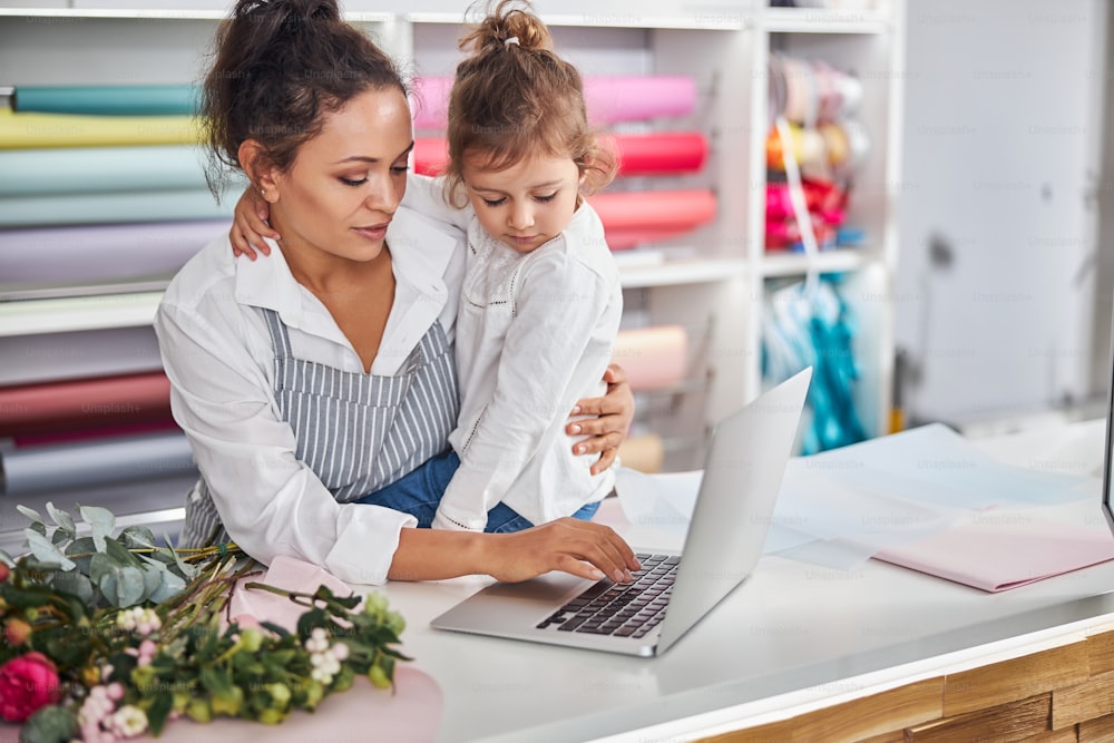 Caring brunette woman holding a little girl in her arms while looking at a laptop screen at a flower shop