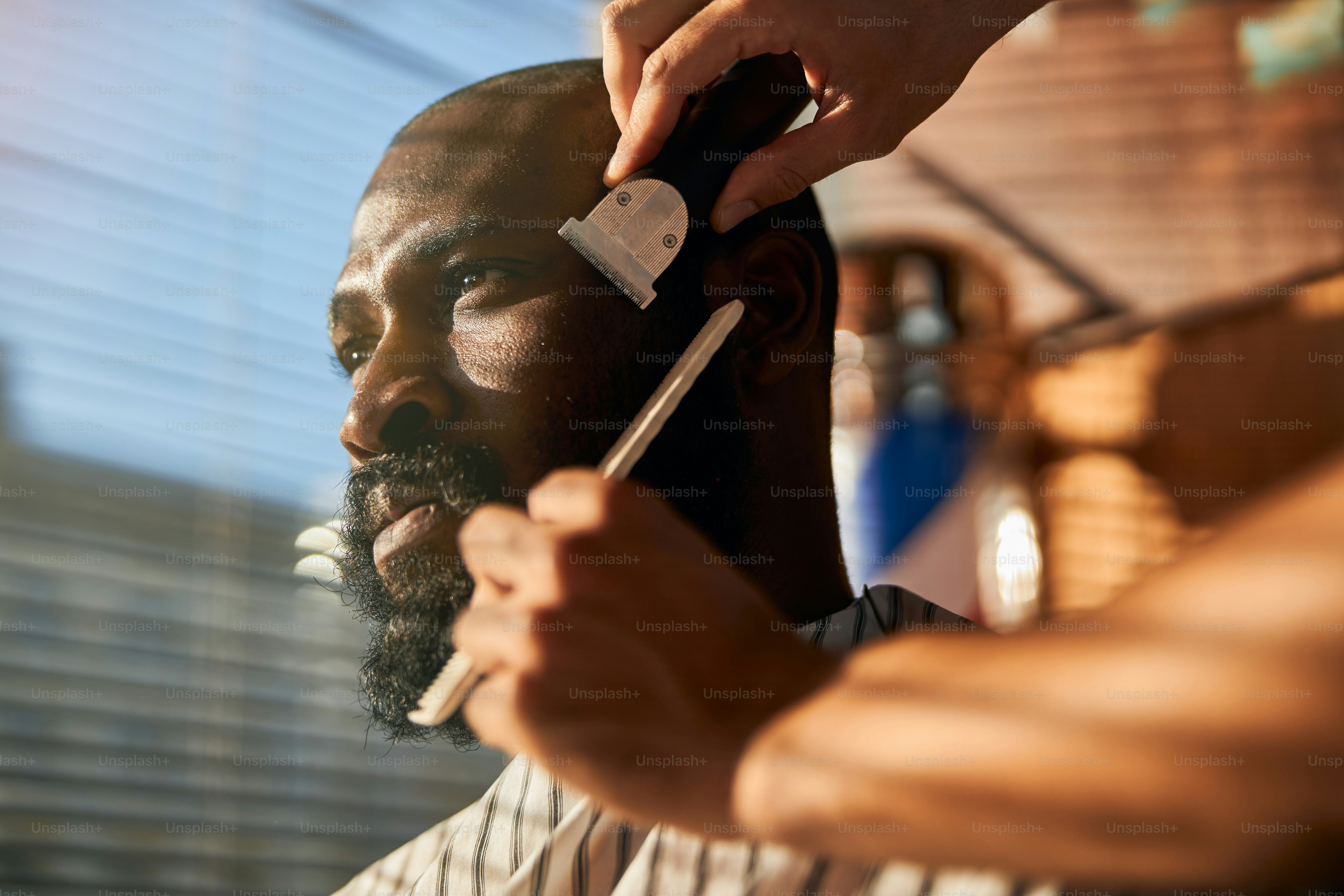 Handsome Afro American man looking away with serious expression while barber removing stubble from his cheek with electric razor