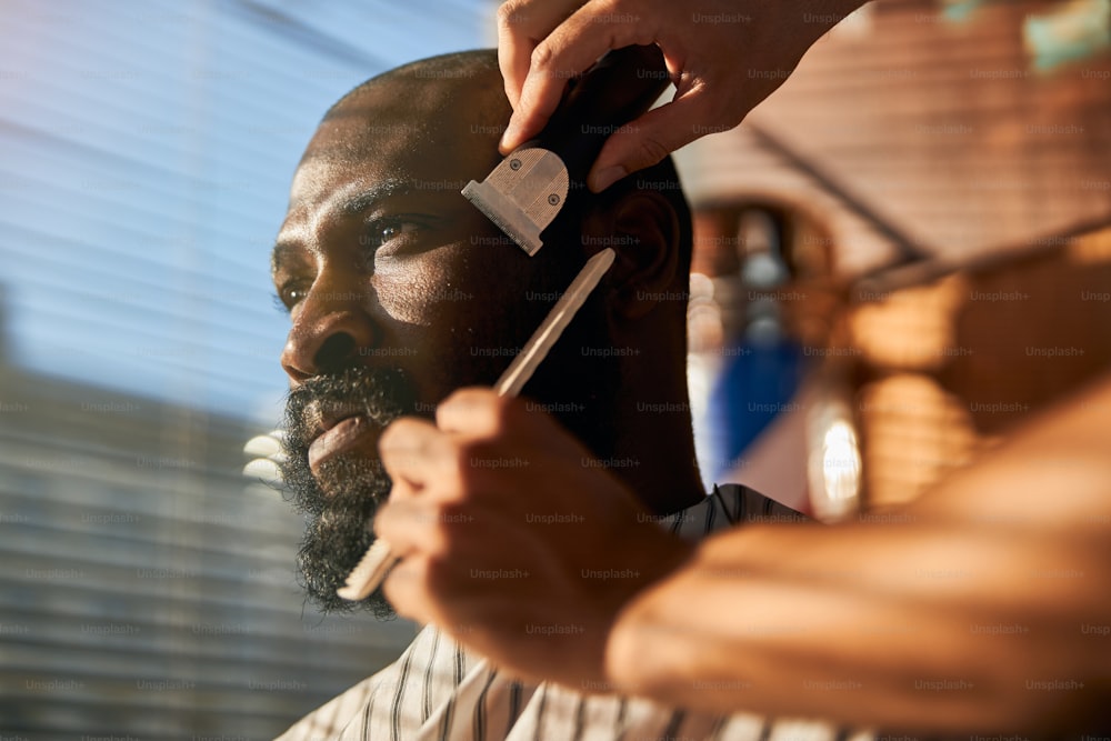 Handsome Afro American man looking away with serious expression while barber removing stubble from his cheek with electric razor