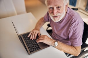 Smiling bearded short-haired disabled Caucasian man sitting in front of his laptop in the wheelchair