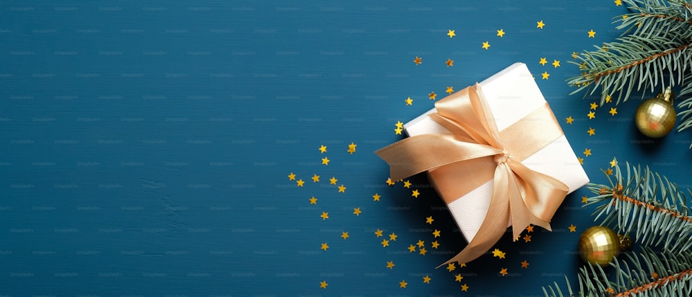 Scatola regalo bianca con fiocco in nastro dorato, palline decorate con rami di abete e coriandoli su sfondo blu. Mockup di banner natalizio, modello di intestazione.