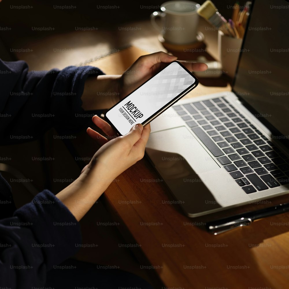Abgeschnittene Aufnahme einer Frau, die das Smartphone hält, während sie mit dem Laptop auf einem Holztisch arbeitet, Clipping-Pfad