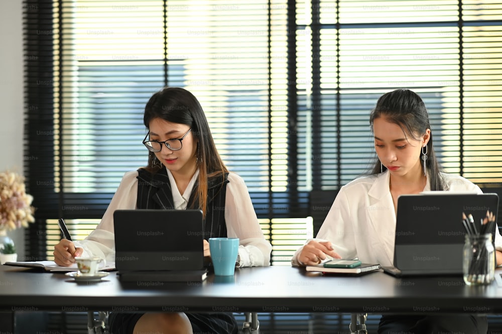 Two young businesswomen working on computer tablet while sitting together in office.