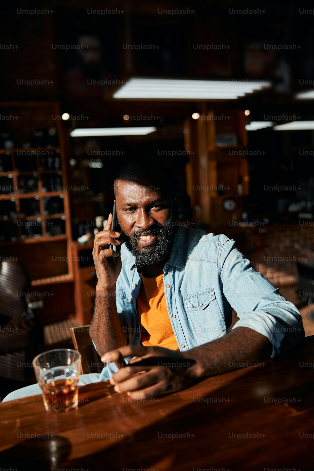 Handsome bearded gentleman having phone conversation and smiling while sitting at the bar counter with glass of whisky