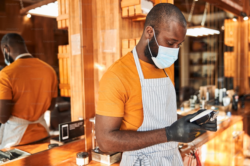 Handsome Afro American man with electric hair trimmer in his hands wearing protective face mask, sterile gloves and apron while standing in barbershop