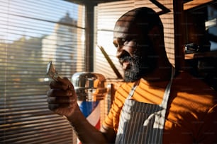 Handsome Afro American young man looking at metal hair clipper and smiling while standing by the window with blinds