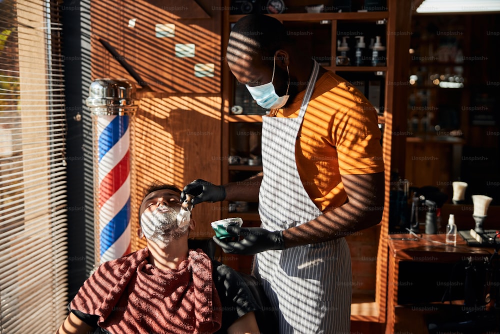 Cabeleireiro masculino afro-americano em máscara facial protetora se preparando para raspar o rosto do homem na barbearia