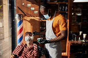 Afroamerikanischer männlicher Friseur in schützender Gesichtsmaske bereitet sich darauf vor, Männergesicht im Friseursalon zu rasieren