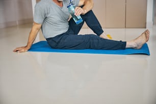 Cropped photo of a barefoot man sitting on a mat and drinking water while taking a break from workout