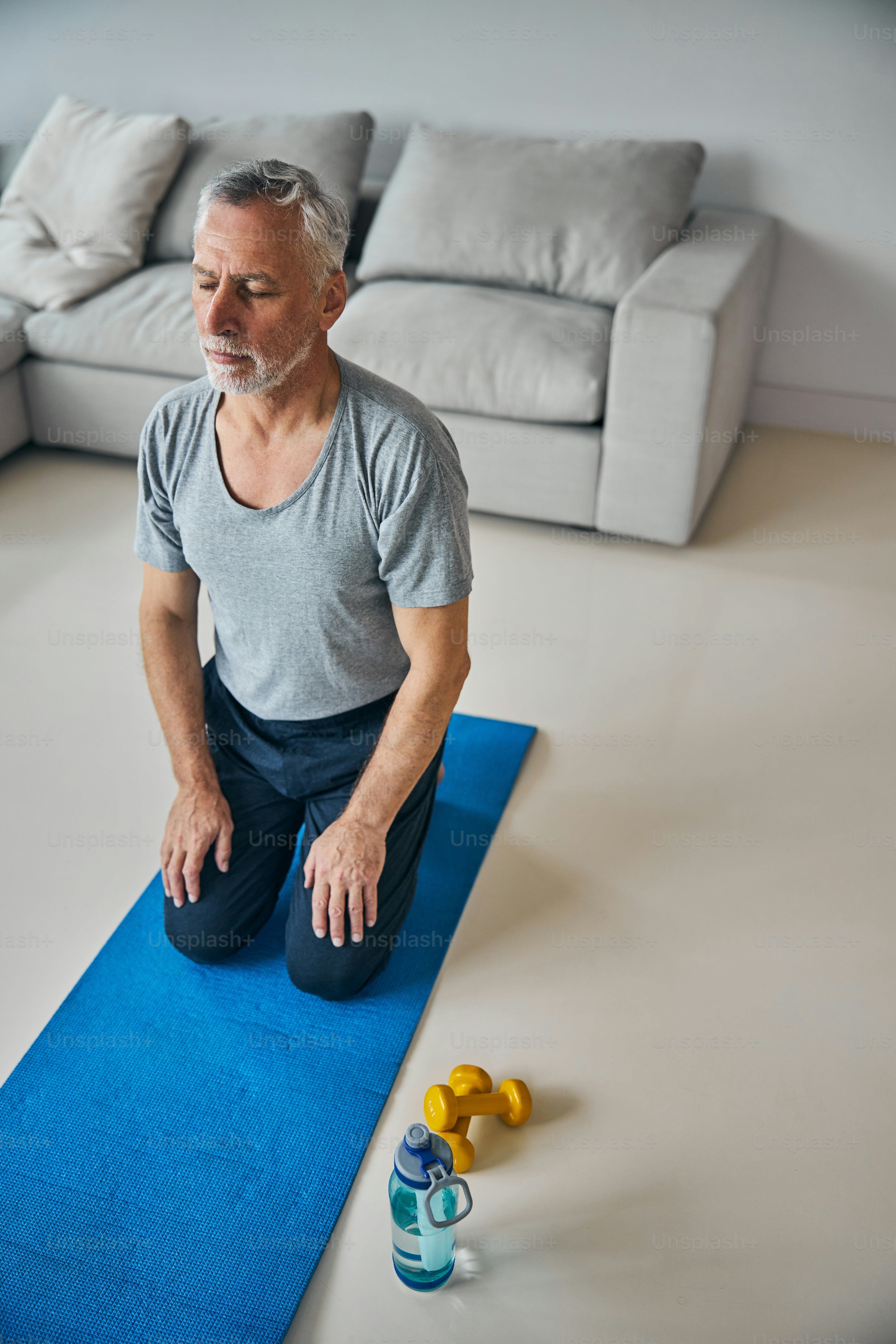 Aged gentleman having his eyes closed while kneeling on a blue yoga mat beside his water bottle