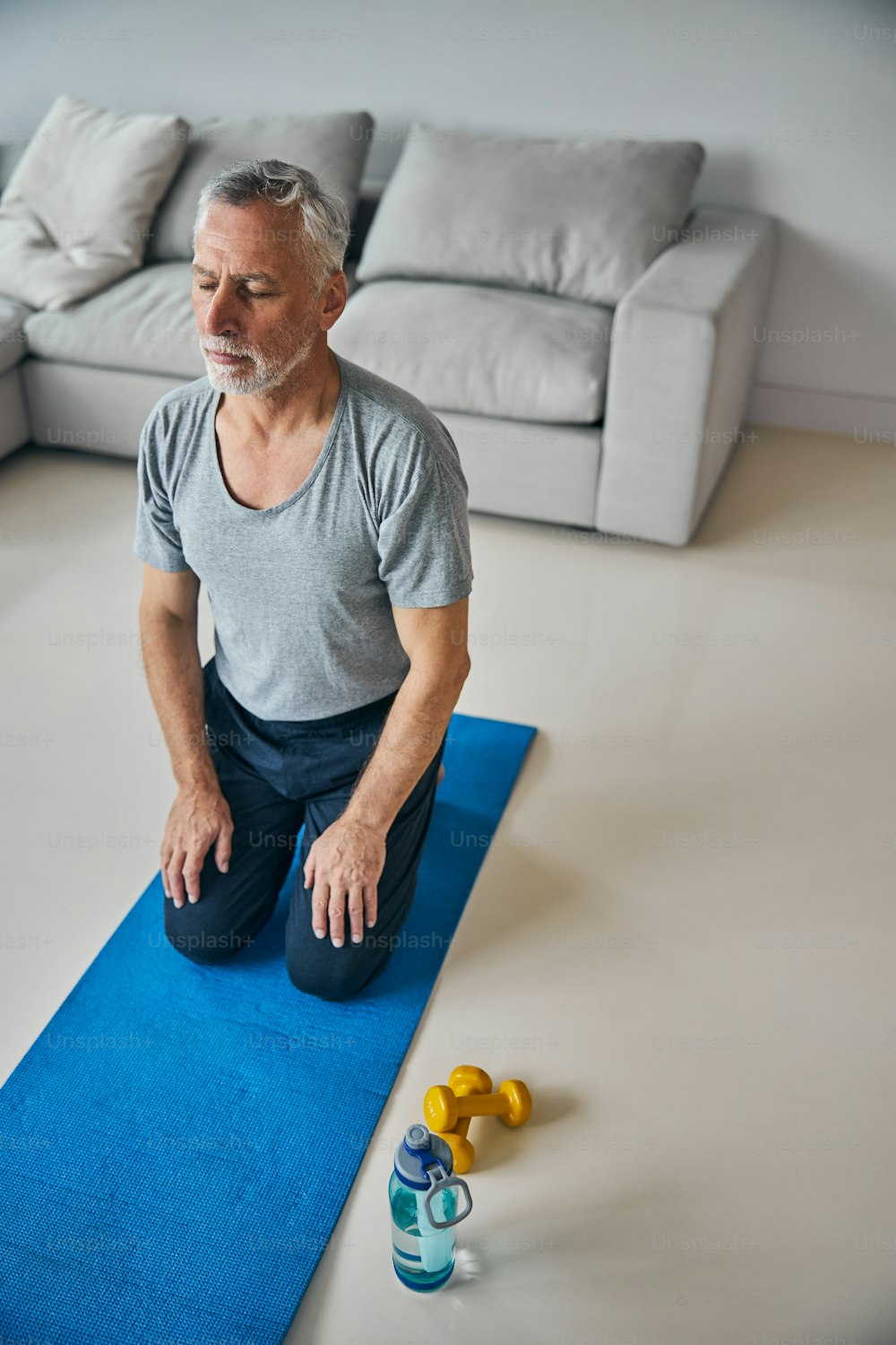 Aged gentleman having his eyes closed while kneeling on a blue yoga mat beside his water bottle
