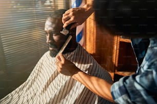 Professioneller Friseur mit Elektrorasierer und Kamm beim Entfernen unerwünschter Haare von der Wange eines jungen Mannes