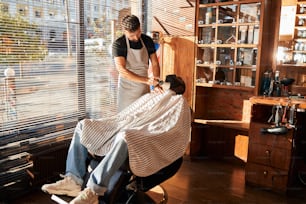Homem afro-americano barbudo sentado na cadeira do salão enquanto cabeleireiro no avental limpando o pescoço com escova de espanador de cabelo