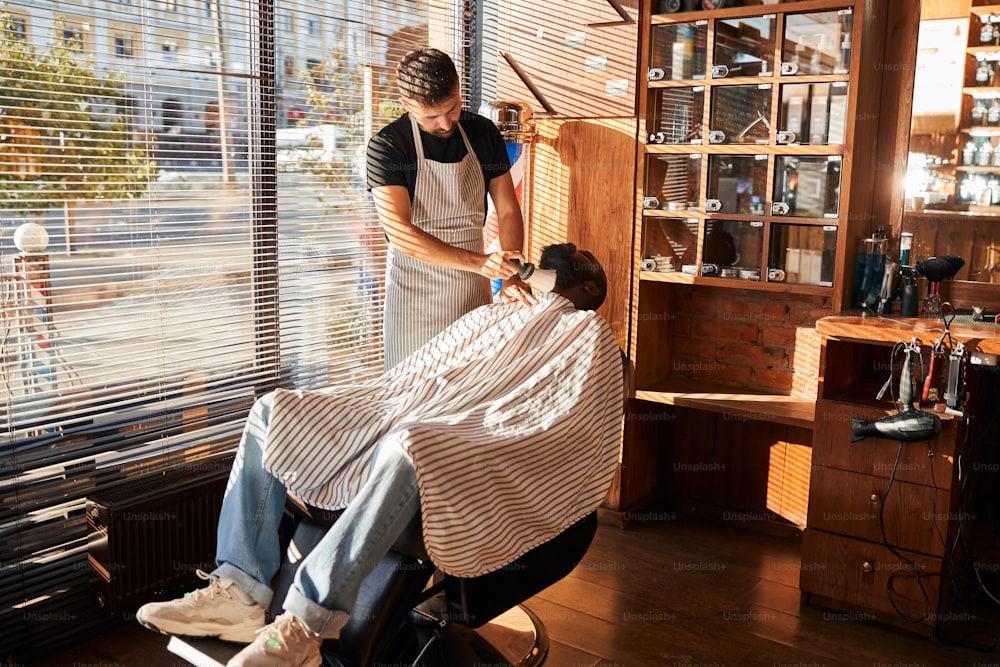 Bearded Afro American man sitting in salon chair while hairdresser in apron cleaning his neck with hair duster brush
