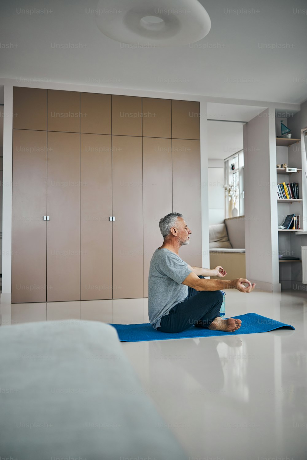 Side-view photo of a senior man meditating while sitting on a workout mat at his appartment