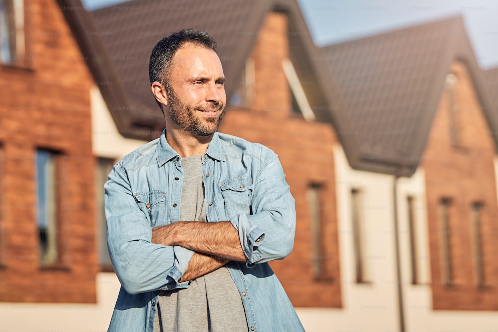 Waist-up photo of a contented man having his arms crossed on chest while standing in front of a new house