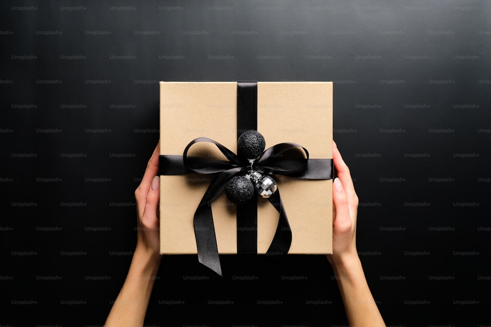 Female hands holding cardboard gift box wrapped black ribbon and Christmas balls over black background. Christmas present, New Year gift box concept.