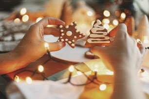 ジンジャーブレッドツリーとスタークッキーを手に、お祝いのライトとクリスマスの飾りが飾られた木製のテーブルの背景にアイシングを施した手。雰囲気のある家族の時間。メリークリスマス