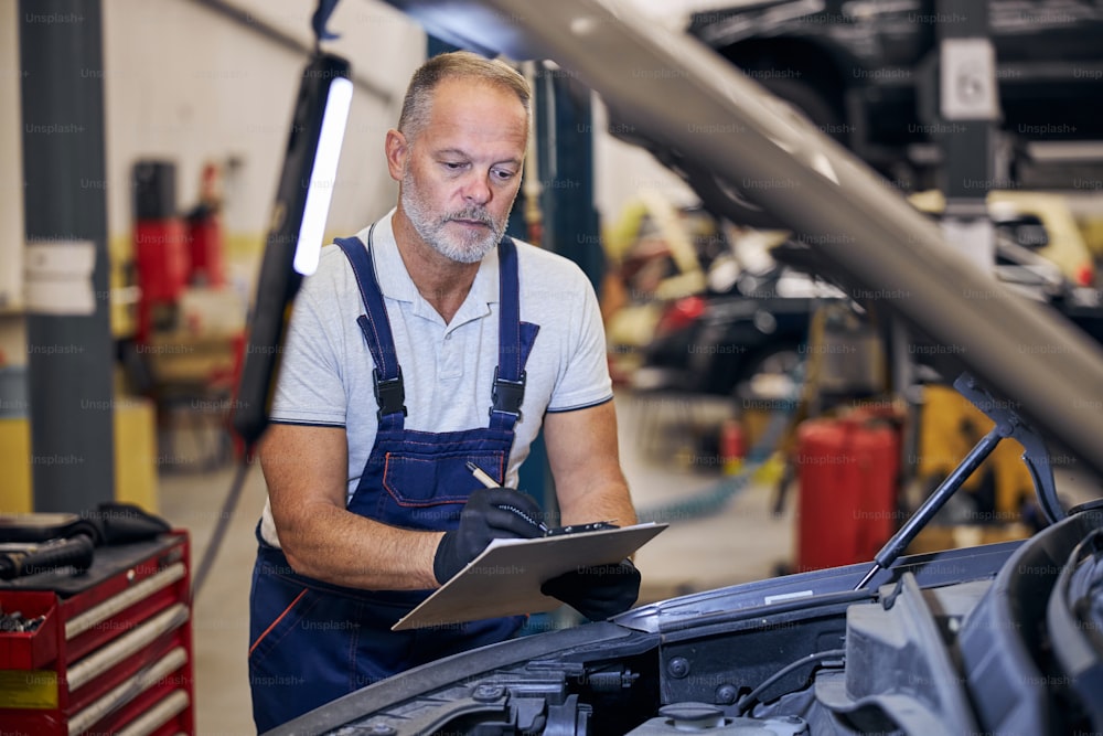 Handsome bearded man in work overalls looking at car engine and writing on clipboard