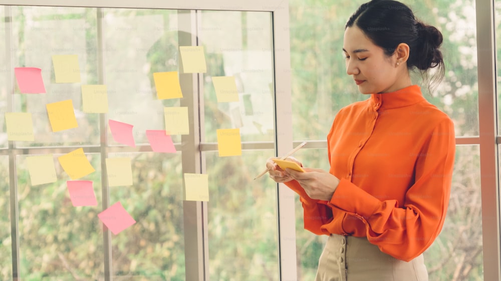 Business people work on project planning board in office and having conversation with coworker friend to analyze project development . They use sticky notes posted on glass wall to make it organized .