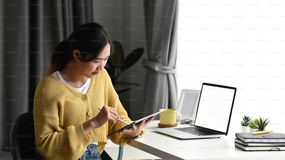Businesswoman in yellow sweater using digital tablet with a stylus pen while sitting in front of blank screen laptop in office.
